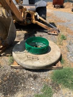 Concrete manhole cover with green ring, wooden supports, near a yellow excavator.