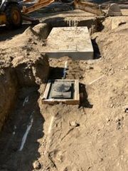 Construction site: Excavated pit with two concrete structures, one with a circular opening. Yellow backhoe visible.
