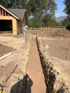 Construction site: trench dug in dirt, with a partly built wood structure in the background. A worker is seen nearby.