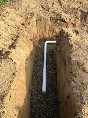 Trench filled with gravel, white PVC pipe extending from trench. Earth walls, outdoor setting.