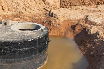A concrete septic tank sits in a shallow excavation filled with murky water.