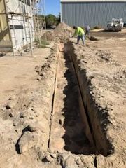 Construction workers digging a trench outdoors near a building. A pile of dirt is visible.