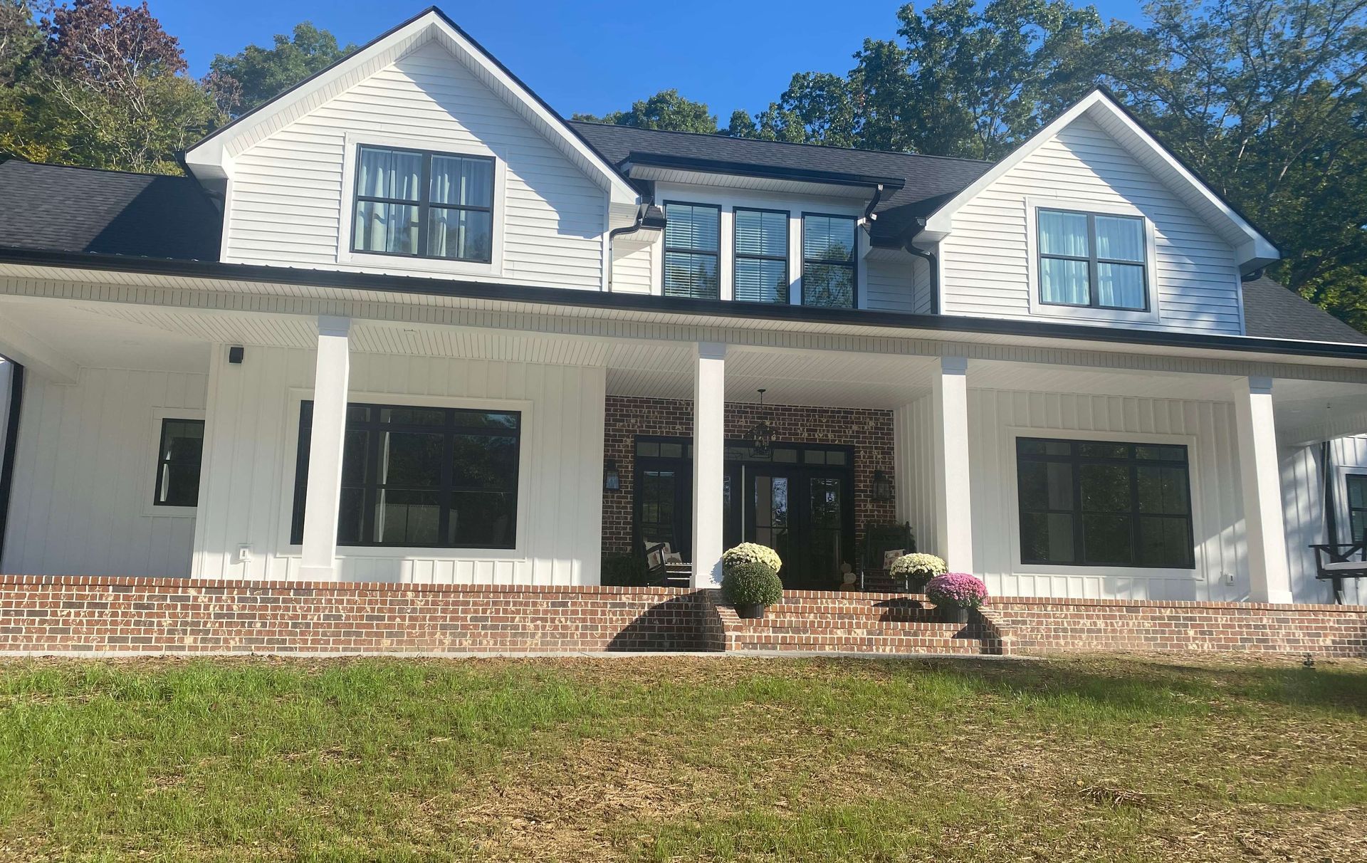 Large white two-story house with a front porch, black shutters, and brick steps on a grassy slope