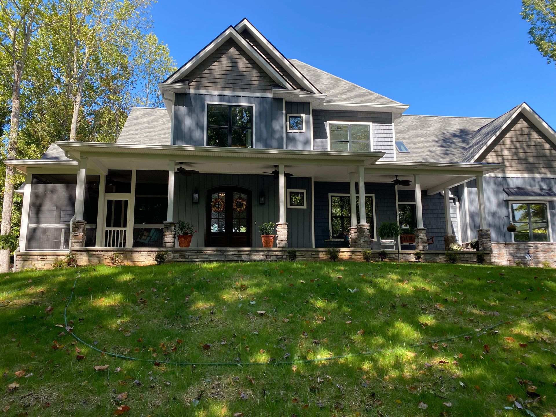 Gray farmhouse with a front porch and green shutters, set on a sunny lawn under a blue sky