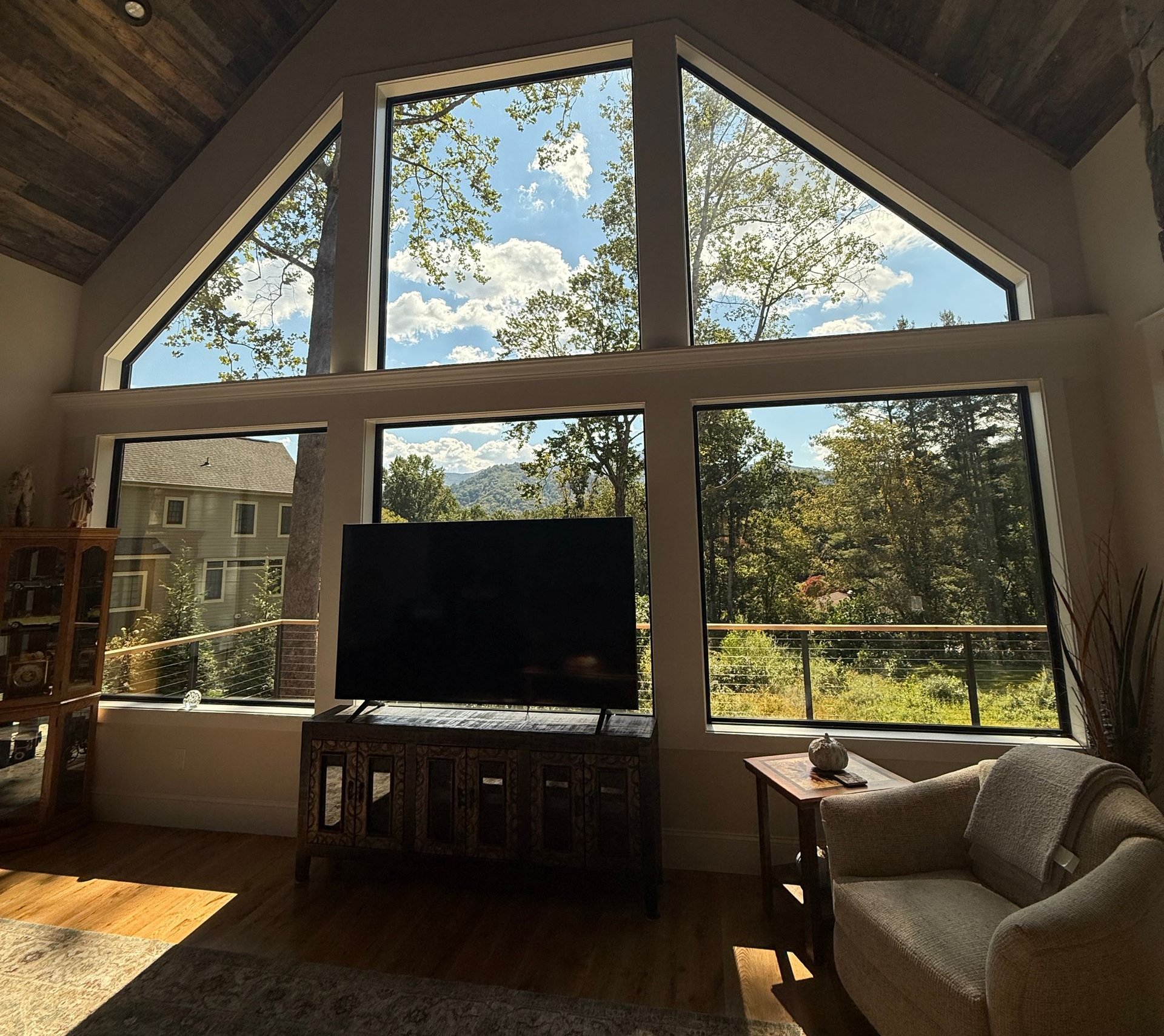 Cozy living room with a large triangular window, TV, armchair, and bright forest view.