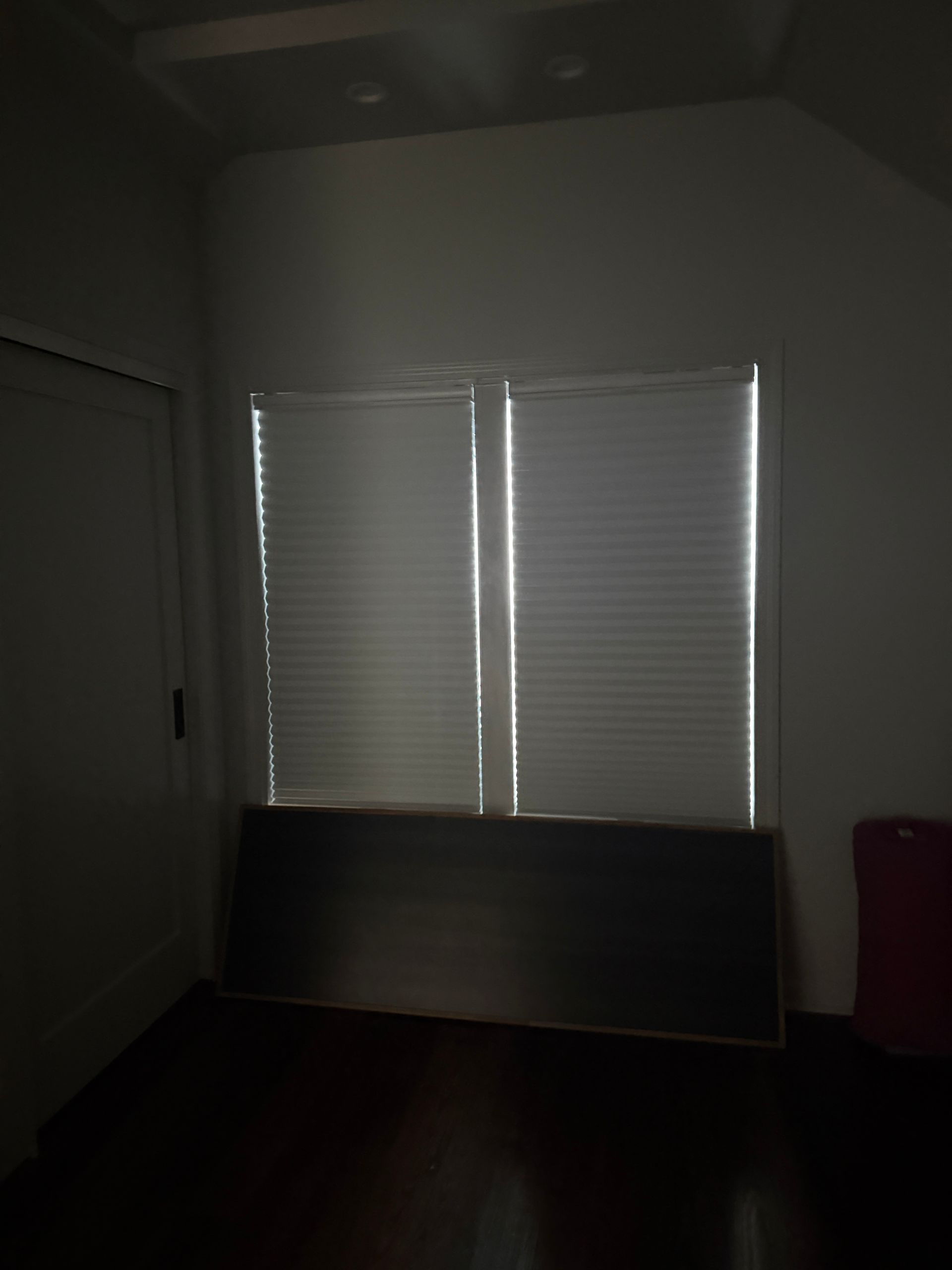 Dim room with white blinds covering a window and a dark board leaning below it.