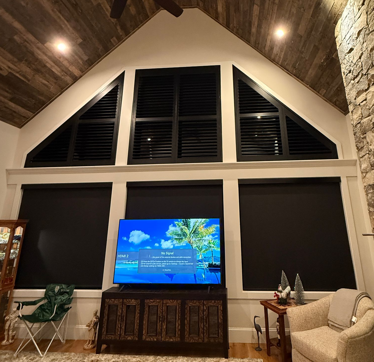 Living room with vaulted wood ceiling, large windows, TV on a wooden console, and a beige chair