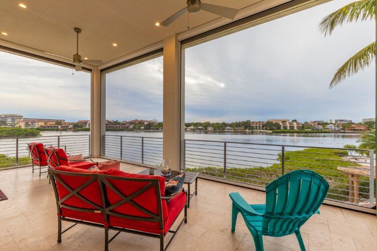 Covered patio with red and teal chairs overlooking a calm waterfront and distant houses.