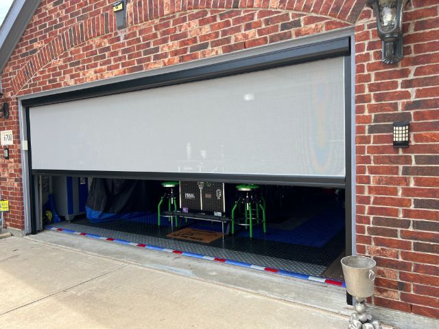 Partially open garage door on a brick house, revealing an indoor pool table area with green stools.