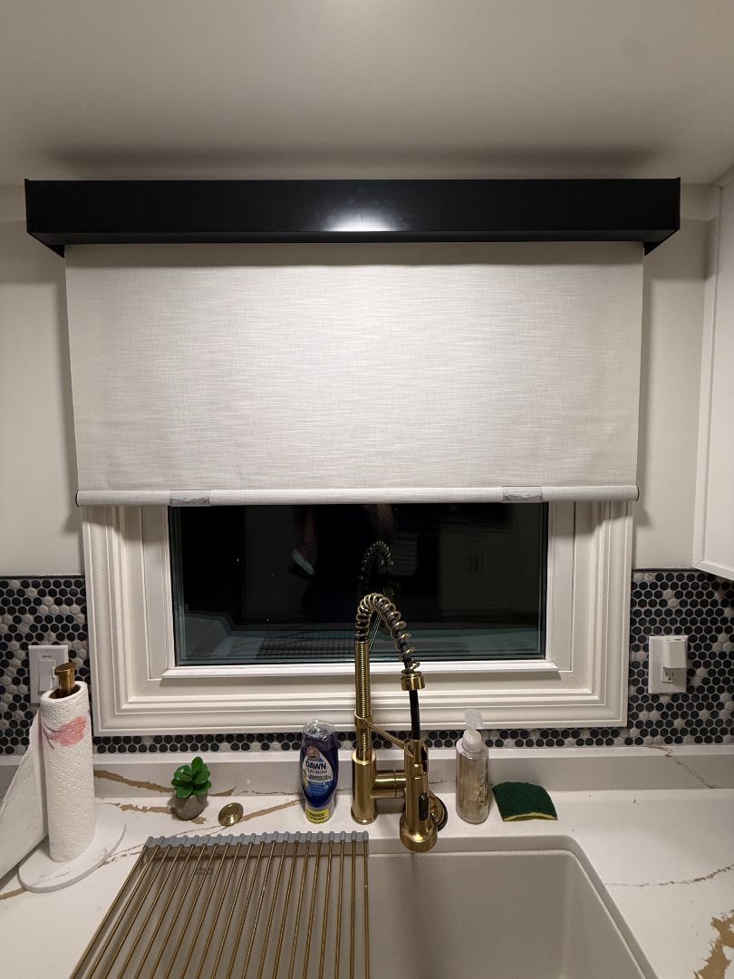Kitchen sink beneath window with white blind, brass faucet, and black window valance.