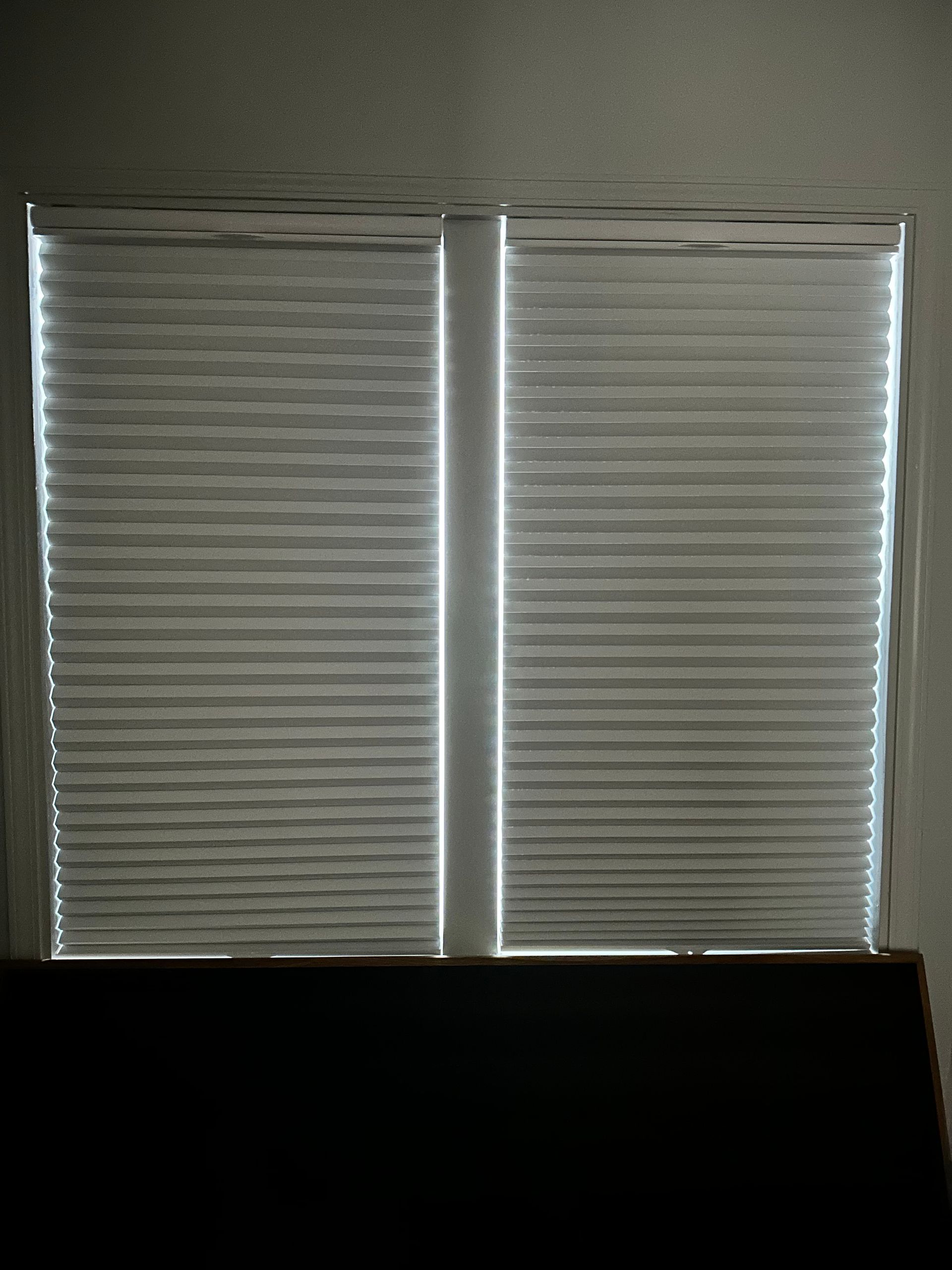 Two white window blinds closed, with light peeking through the slats.