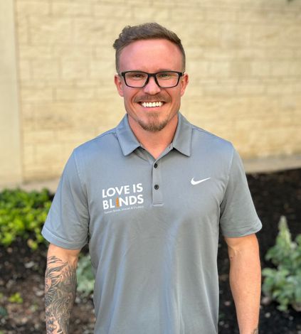 Smiling person in a gray polo shirt standing outdoors against a beige wall.
