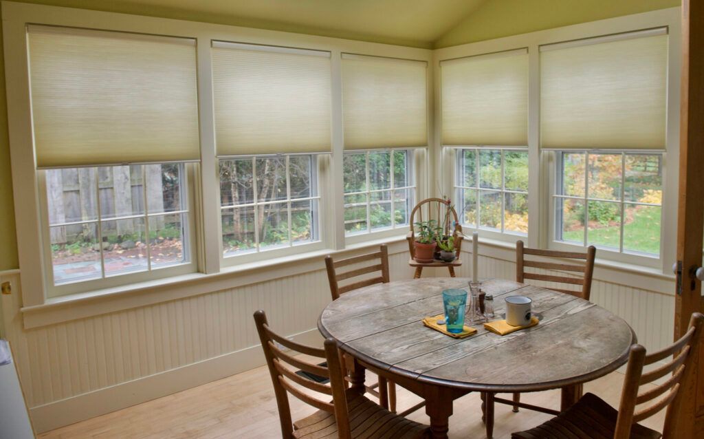 Sunlit dining nook with round wooden table, chairs, and large windows with blinds