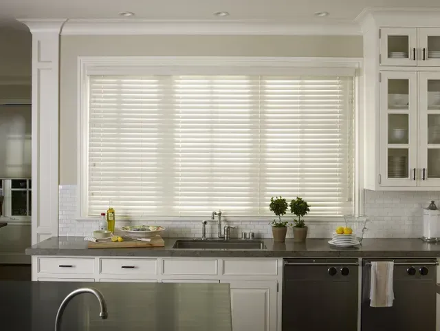 Bright white kitchen with large window over a sink, marble counters, and dark dishwasher below