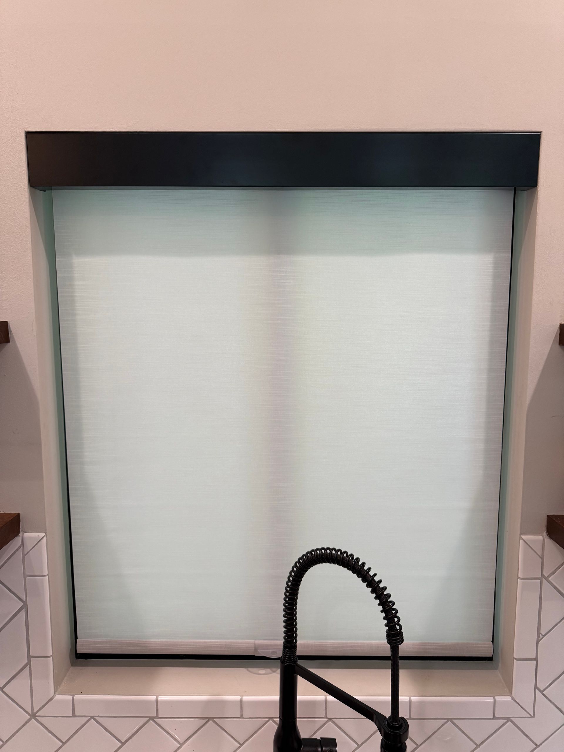 Closed white window blind with black valance above a black faucet in the foreground