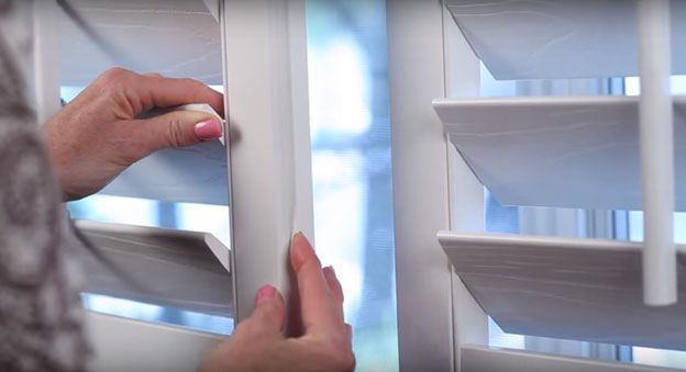 Hands opening white window shutters with slats in a bright room
