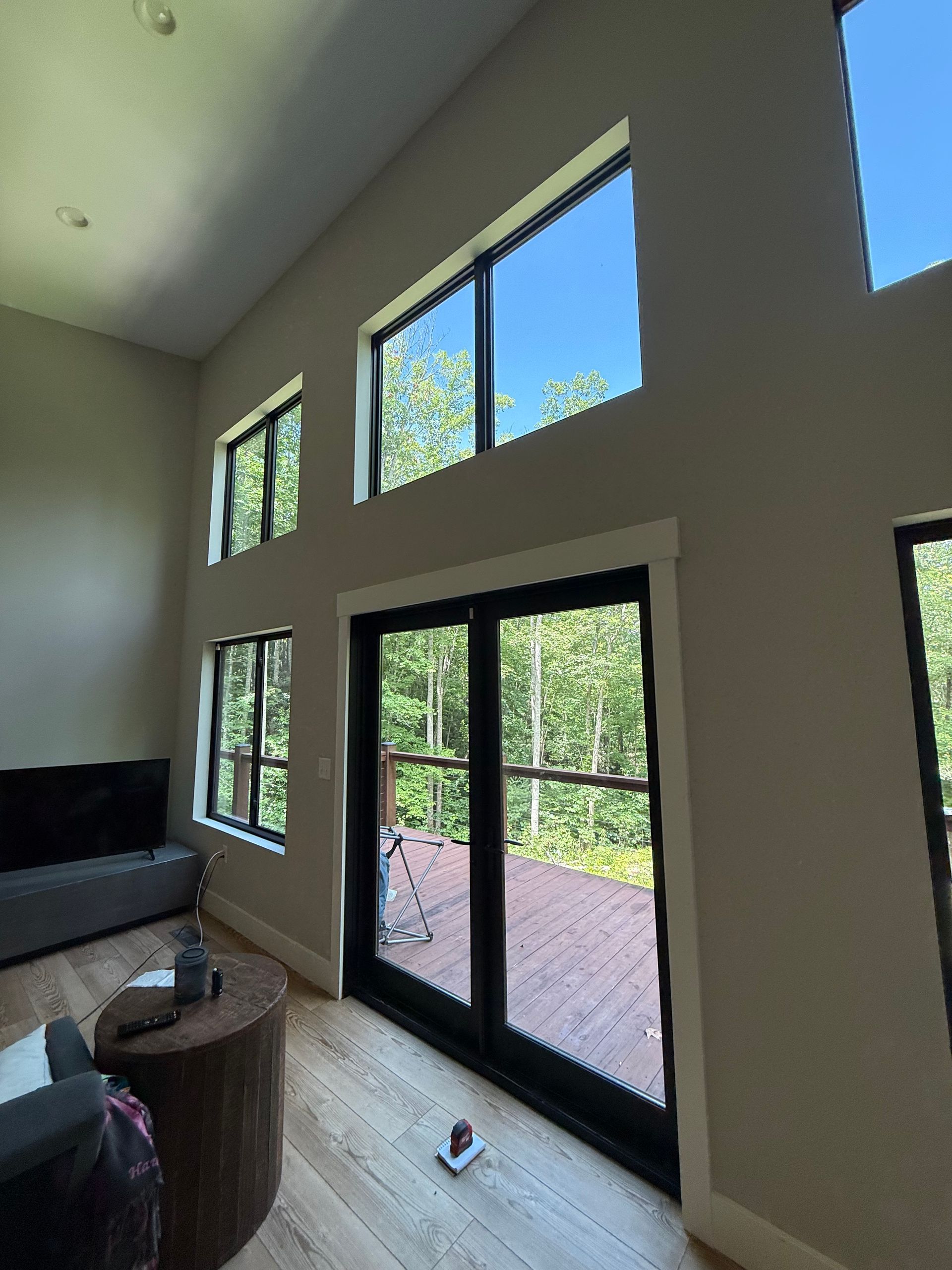 Bright living room with tall windows and glass doors opening to a sunlit deck and green trees