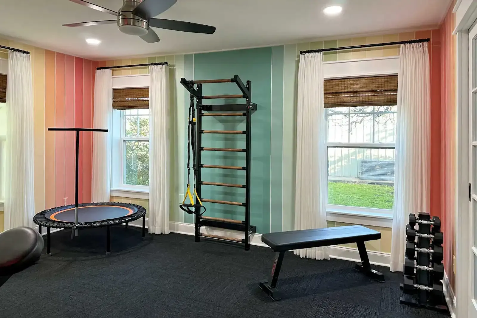 Home gym with trampoline, wall ladder, bench, dumbbells, and large windows with curtains.