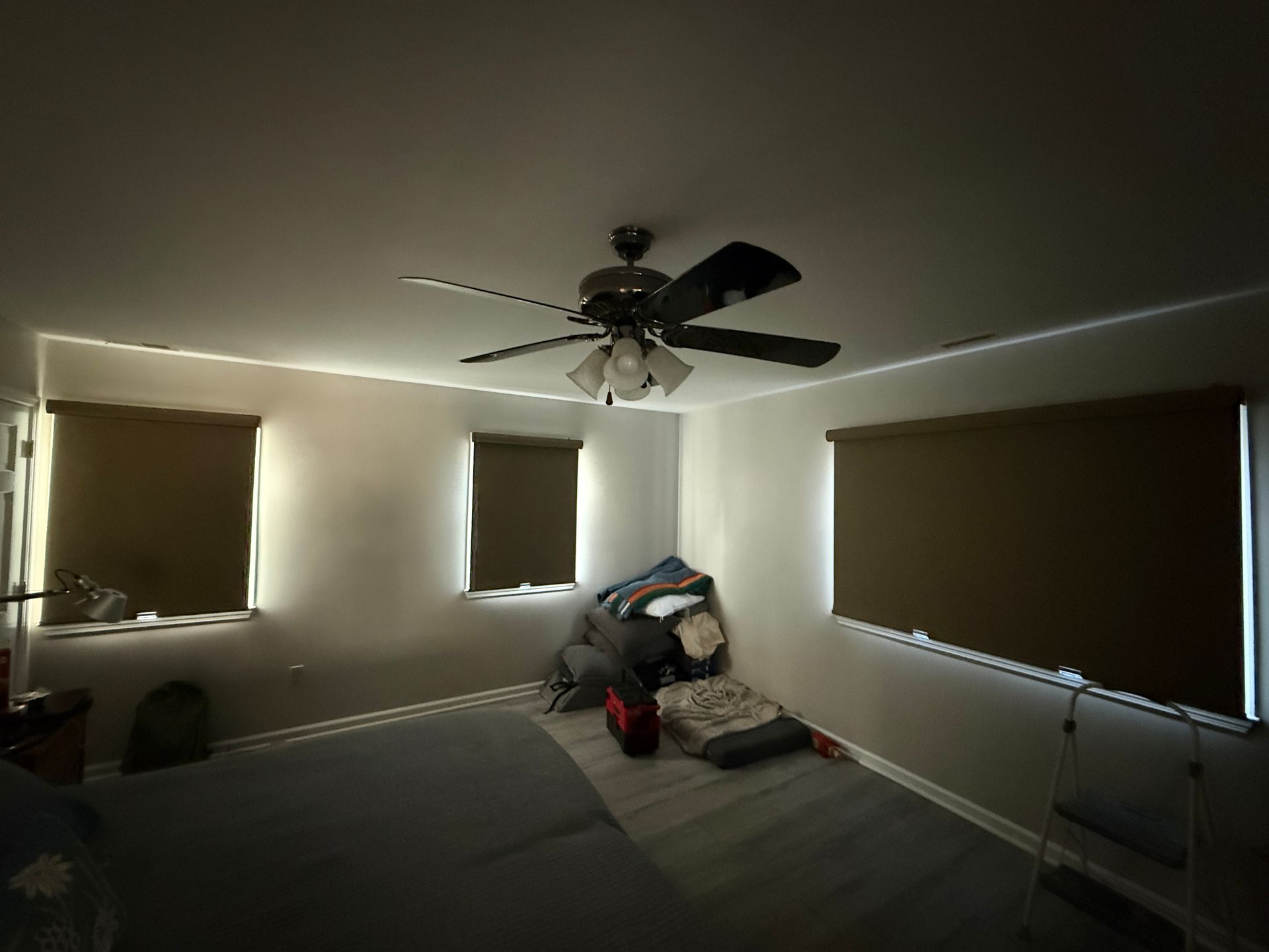 Dim bedroom with a ceiling fan, three windows, and a pile of clothes on the bed.