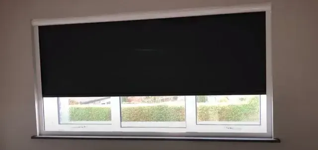 Window with a black roller blind pulled down, leaving a narrow view of the outside.