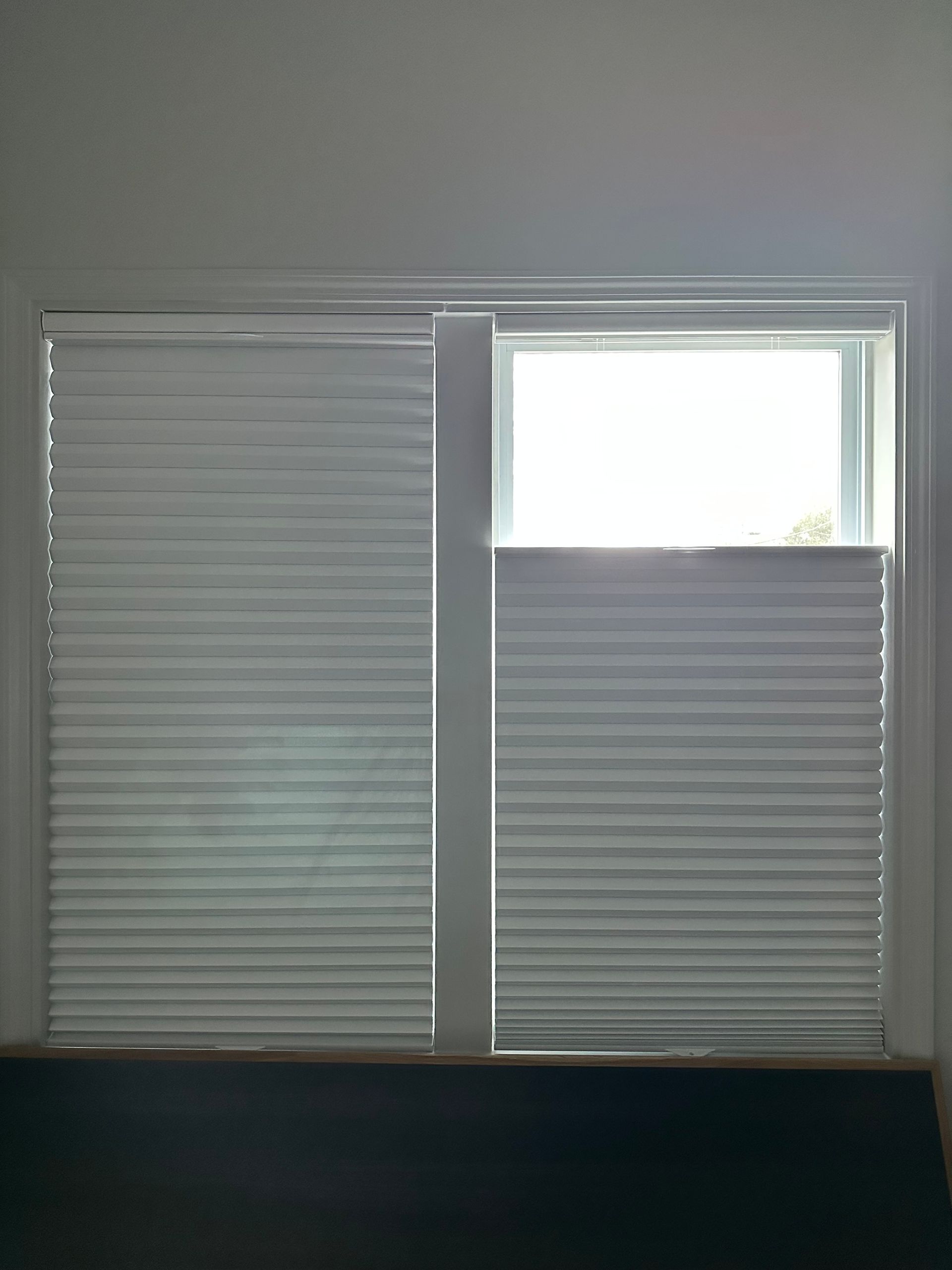 Two white window blinds, one partly open revealing bright daylight above the other.
