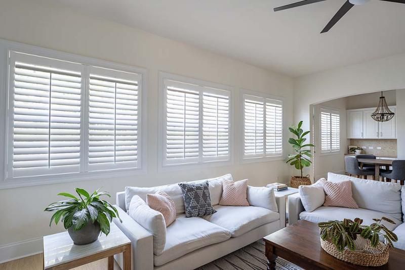 Bright living room with white shutters, cream sofas, potted plants, and a dining area in the background