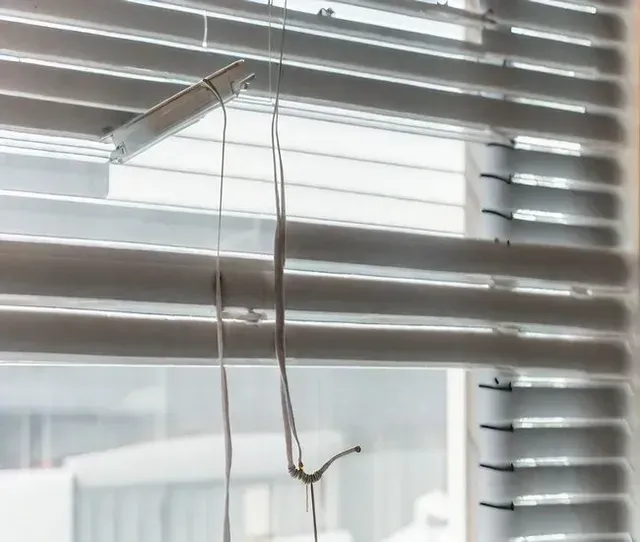 A hanging cord beside white window blinds with bright daylight filtering through.