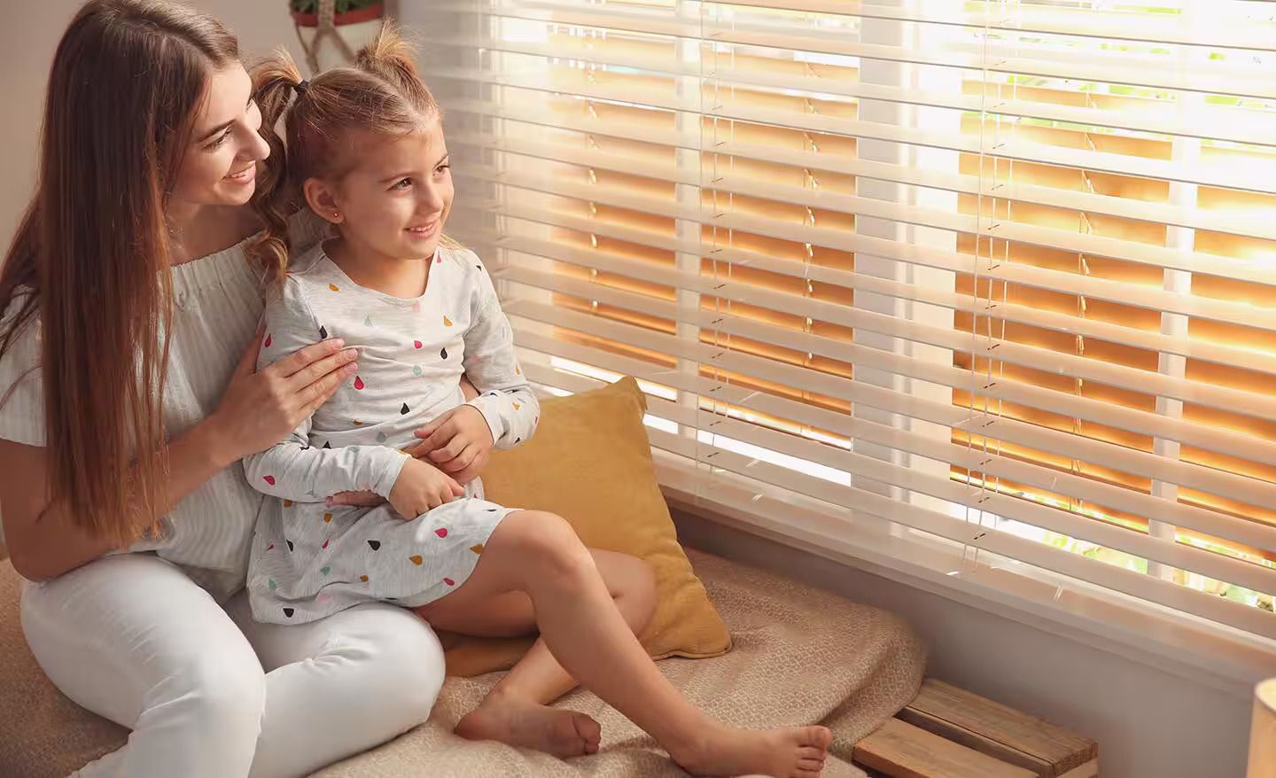 Two people sitting by a window, looking out through blinds in warm sunlight.