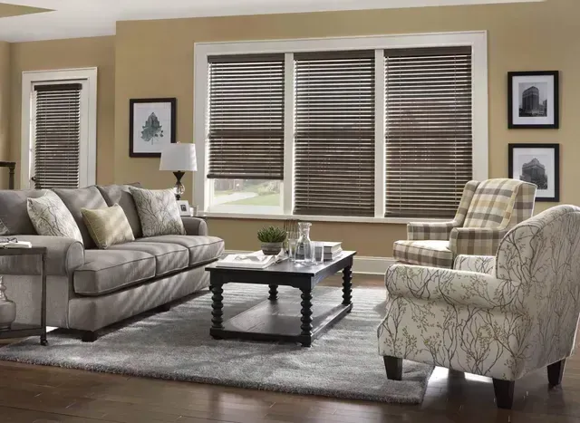 Bright living room with beige sofas, patterned armchair, coffee table, and large windows with blinds.