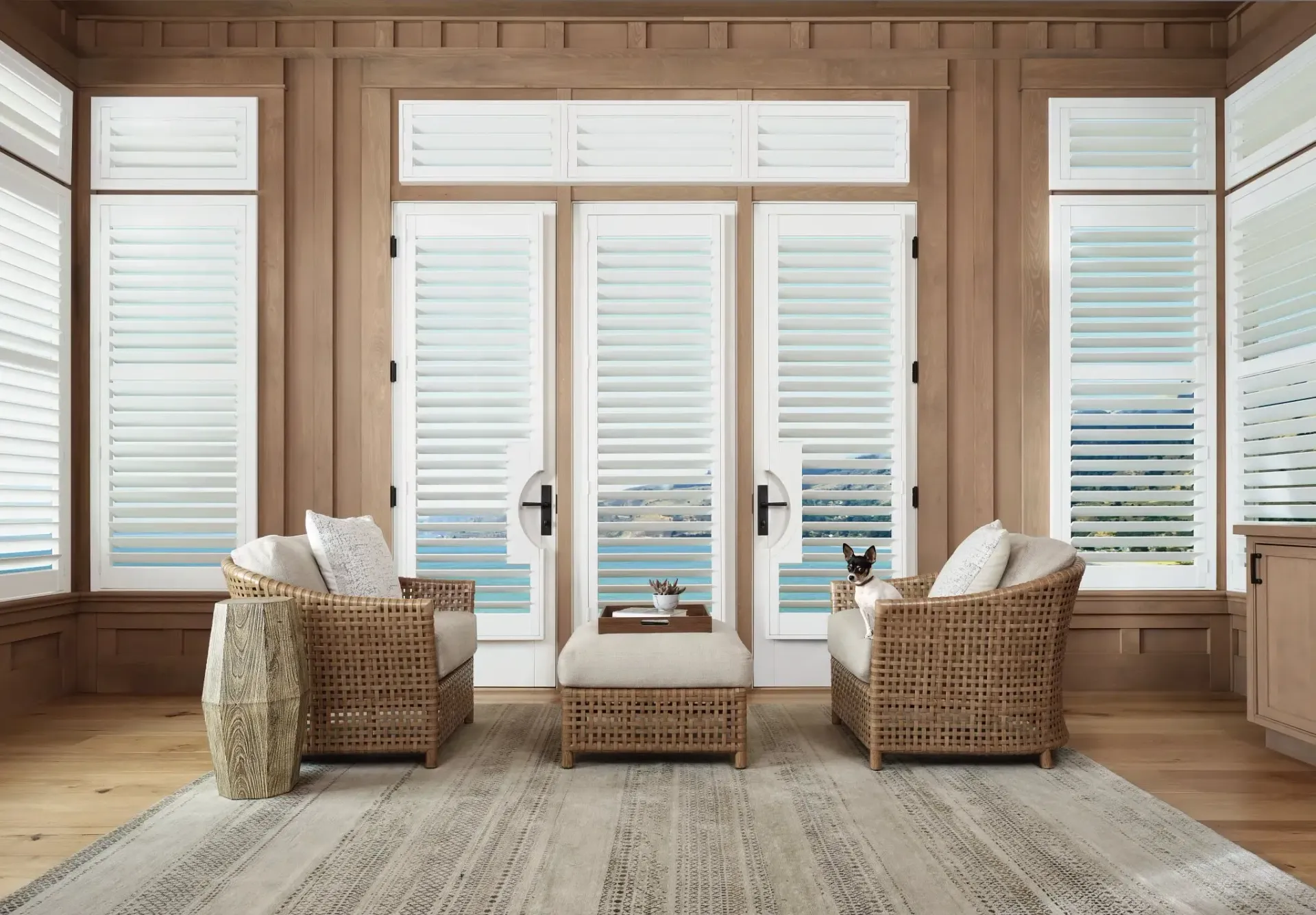 Bright sunroom with wicker chairs, ottoman, and sheer curtains overlooking a deck and water.