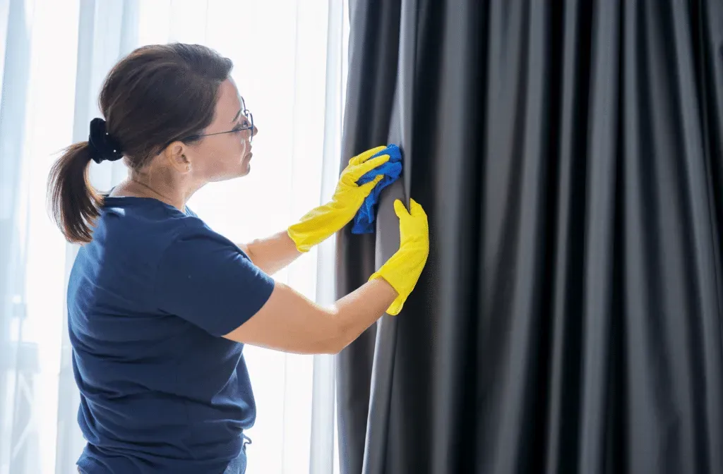 Person in blue shirt and yellow gloves cleaning a dark curtain by a window