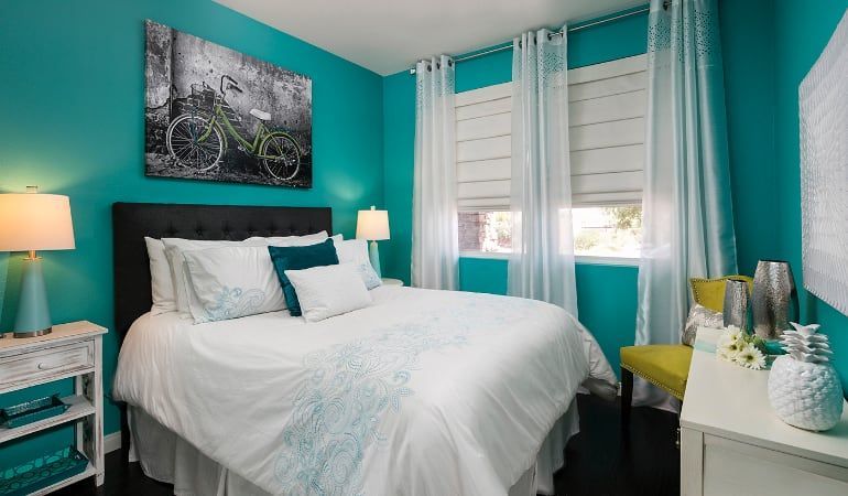 Bright teal bedroom with white bed, windows, lamp, and dresser decor