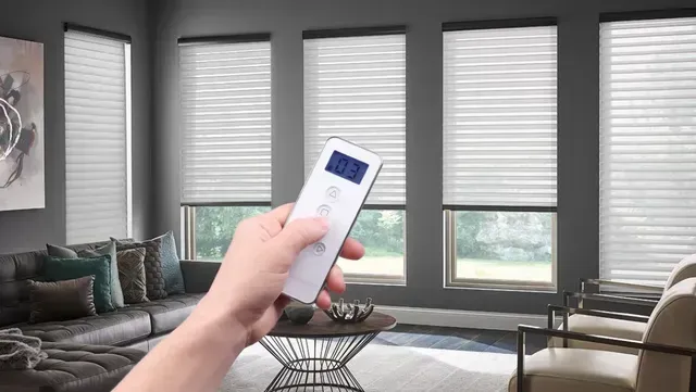 Hand holding a white remote pointed at living room window blinds with filtered daylight