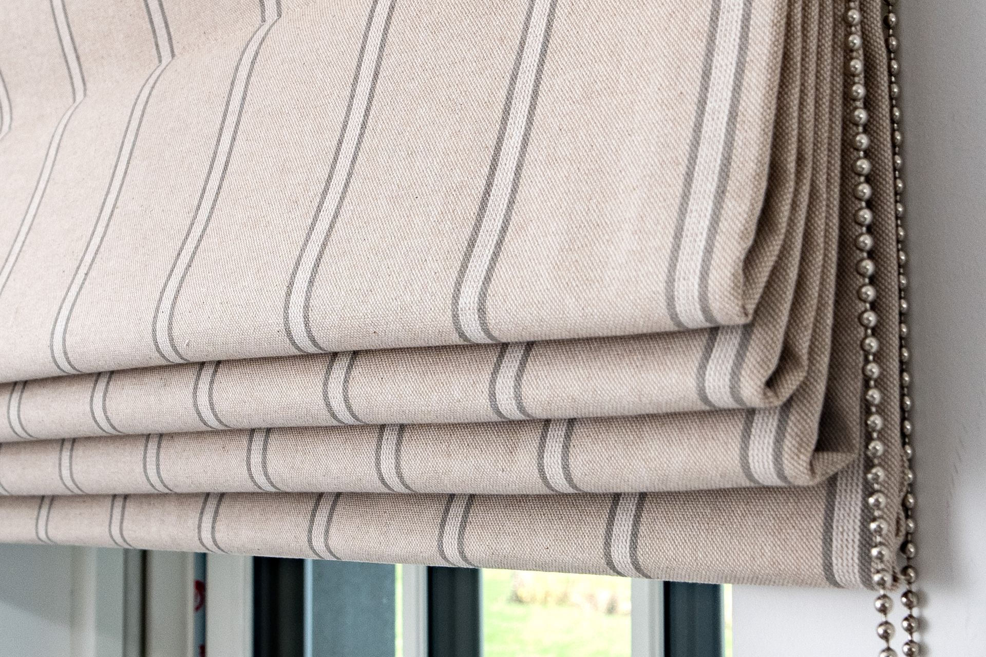 Beige striped roman blinds stacked at a window with hanging bead chains