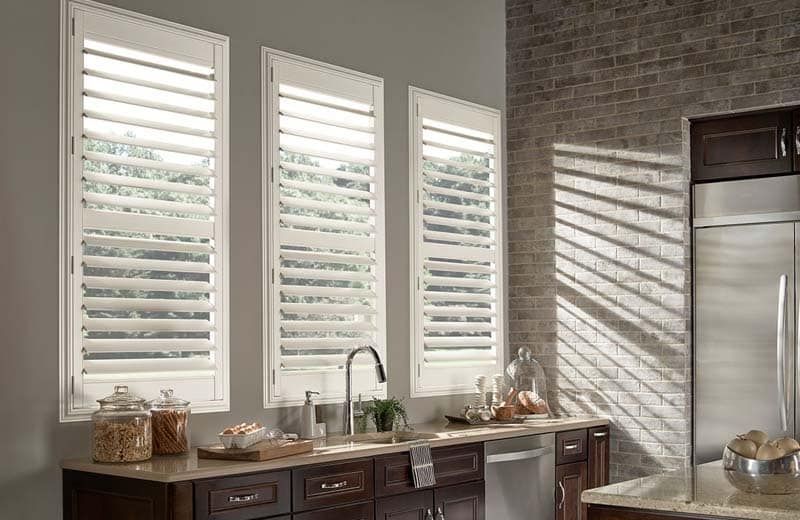 Bright kitchen with tall windows, white shutters, dark cabinets, and sunlight casting striped shadows.