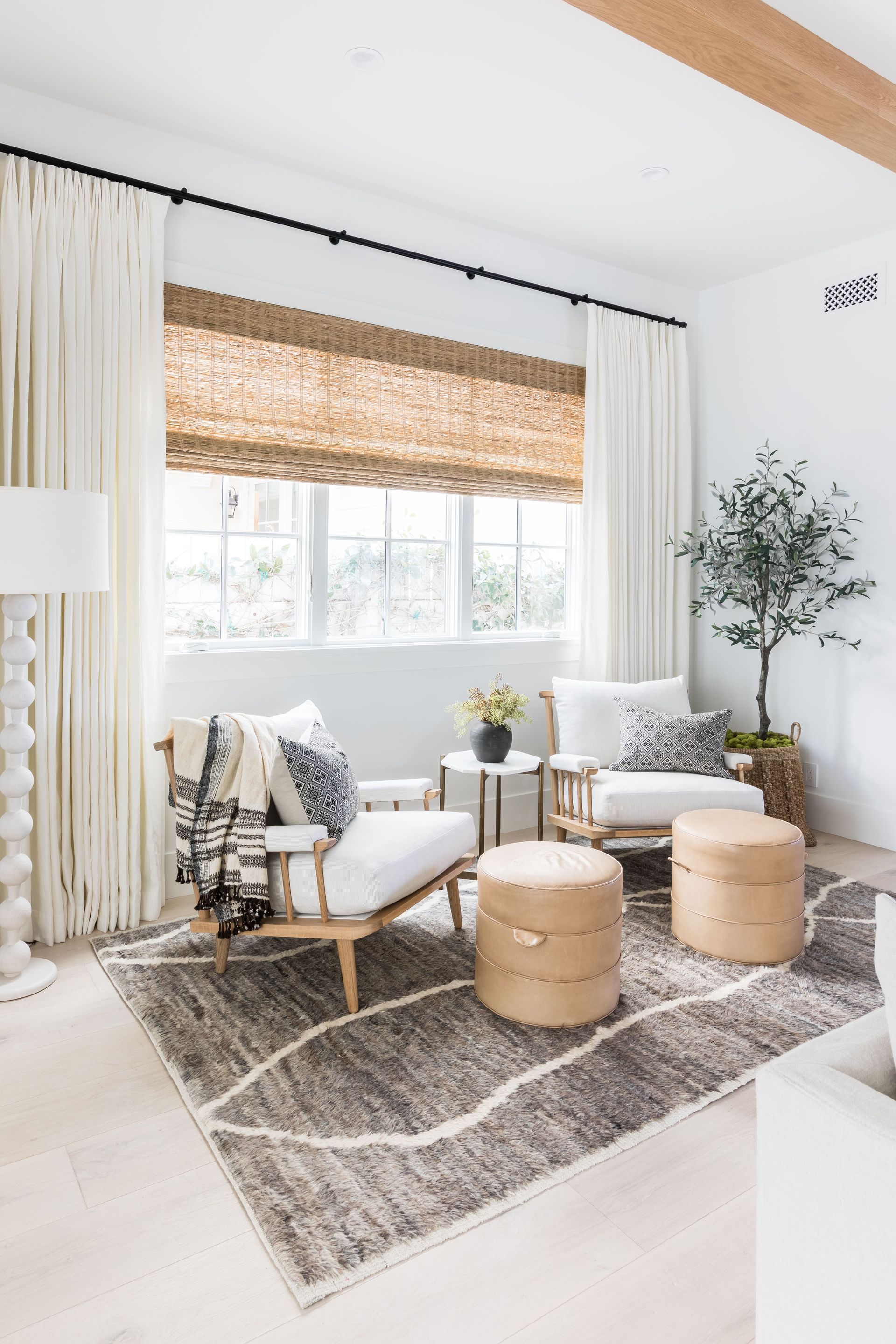Bright living room with white chairs, round ottomans, woven shade, and large window on a patterned rug