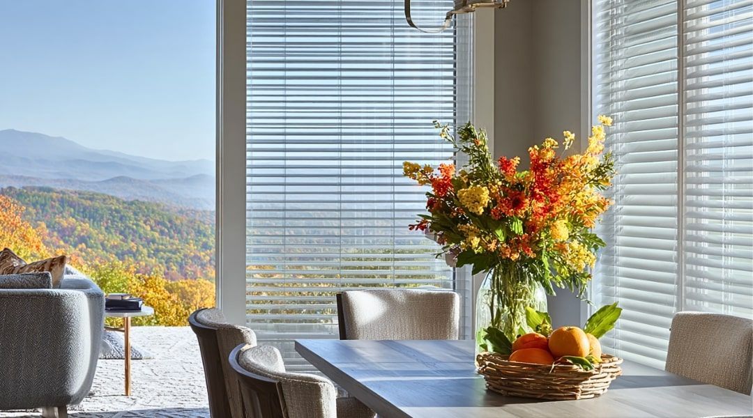 Bright dining room with a table, flowers, fruit bowl, and mountain view through large windows