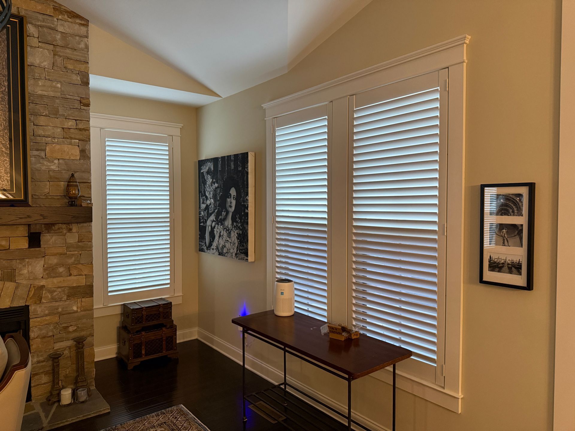 Living room corner with large windows, white blinds, and a dark wood console table with blue accent light