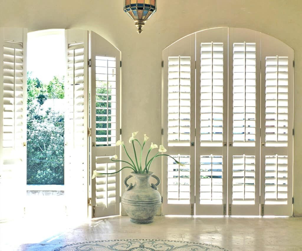 Bright room with open white shutters, arched windows, and a vase of flowers on the floor
