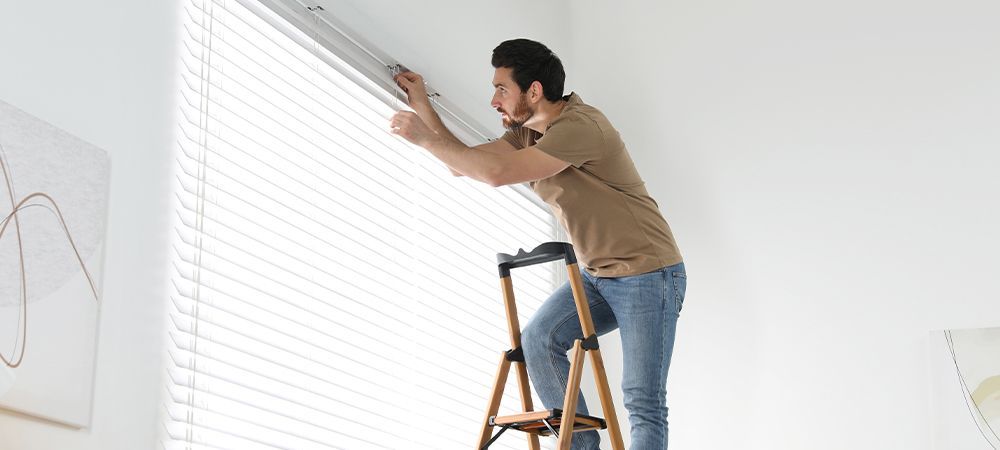 Man on a ladder installing blinds on a window in a bright room