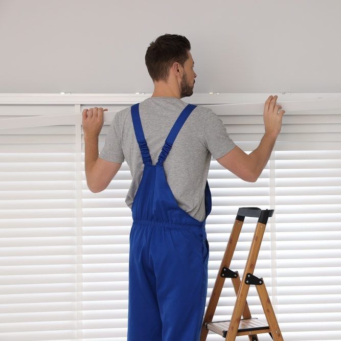 Man in blue overalls adjusting white blinds beside a wooden step ladder