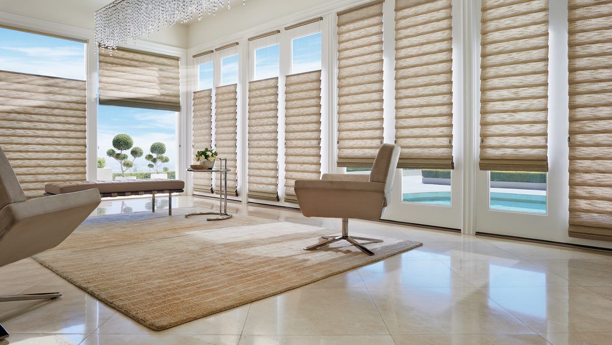 Bright modern living room with beige chairs, woven rug, and tall windows with shutters overlooking water.