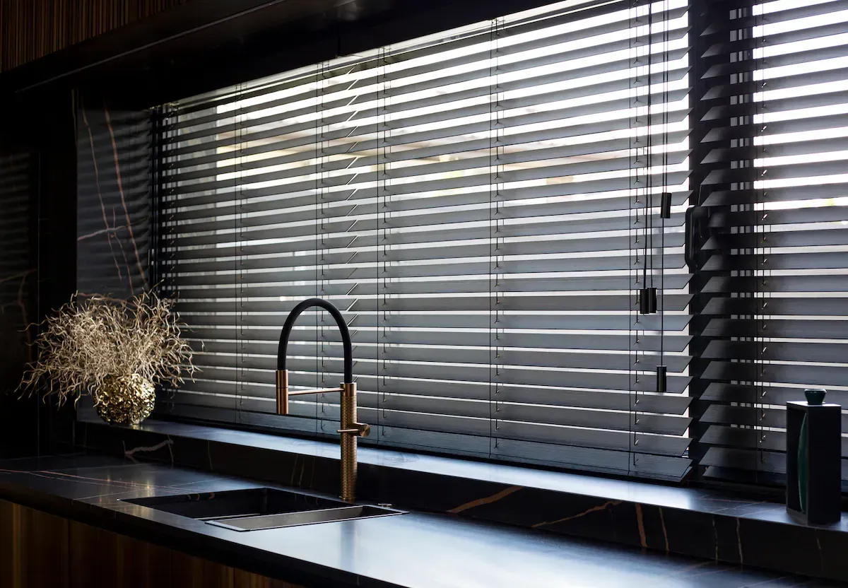 Modern kitchen sink under wide window with gray blinds and a vase of dried flowers