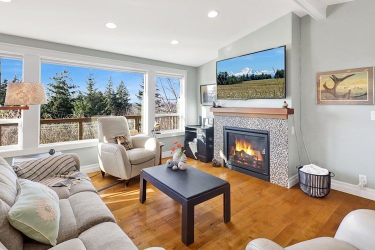 Bright living room with large windows, gray sofa, armchair, fireplace, and wall-mounted TV