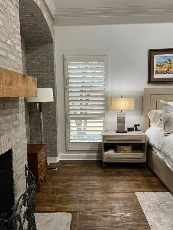 Modern bedroom with a brick fireplace, white shutters, bedside table lamp, and neatly made bed