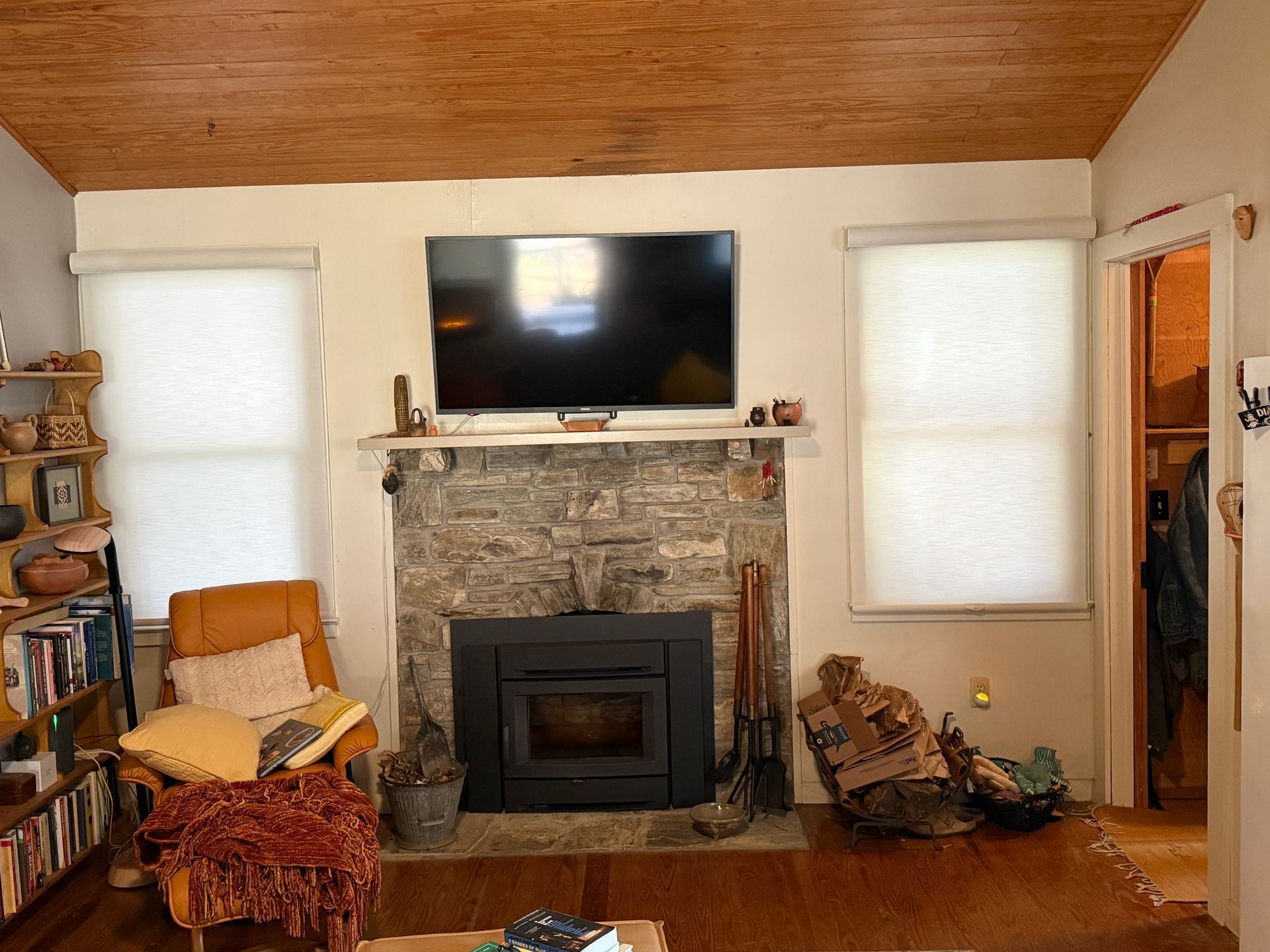 Living room with stone fireplace, TV, and two windows with blinds. Wooden ceiling and hardwood floor.