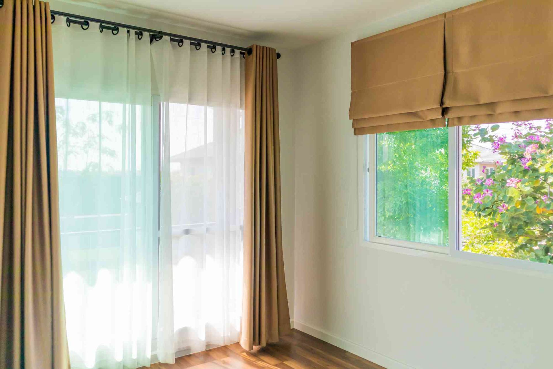 Bright empty room with sheer white curtains, tan drapes, and a window showing greenery outside