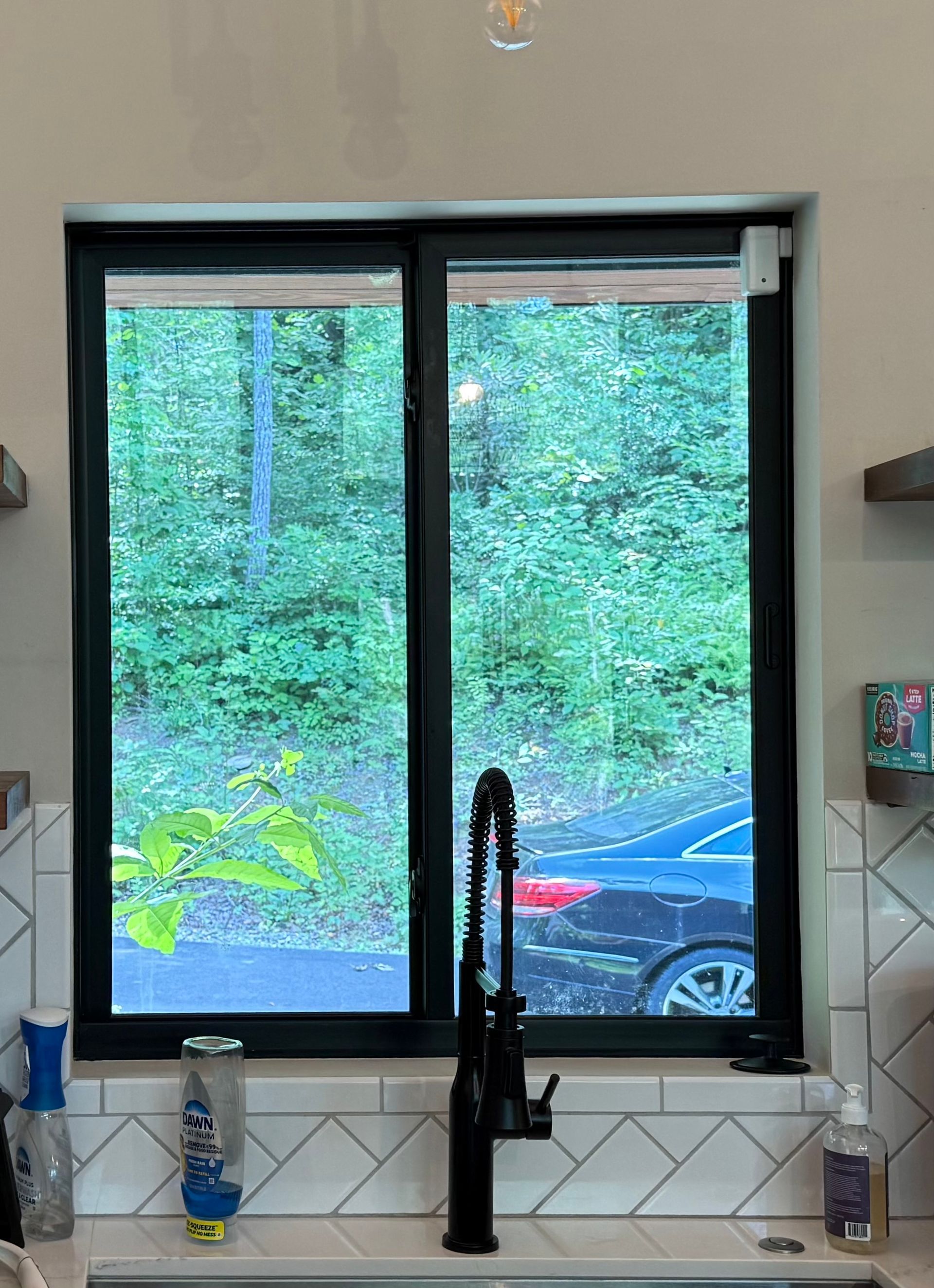 Kitchen window over sink with green trees outside, black faucet centered, and parked car visible through glass