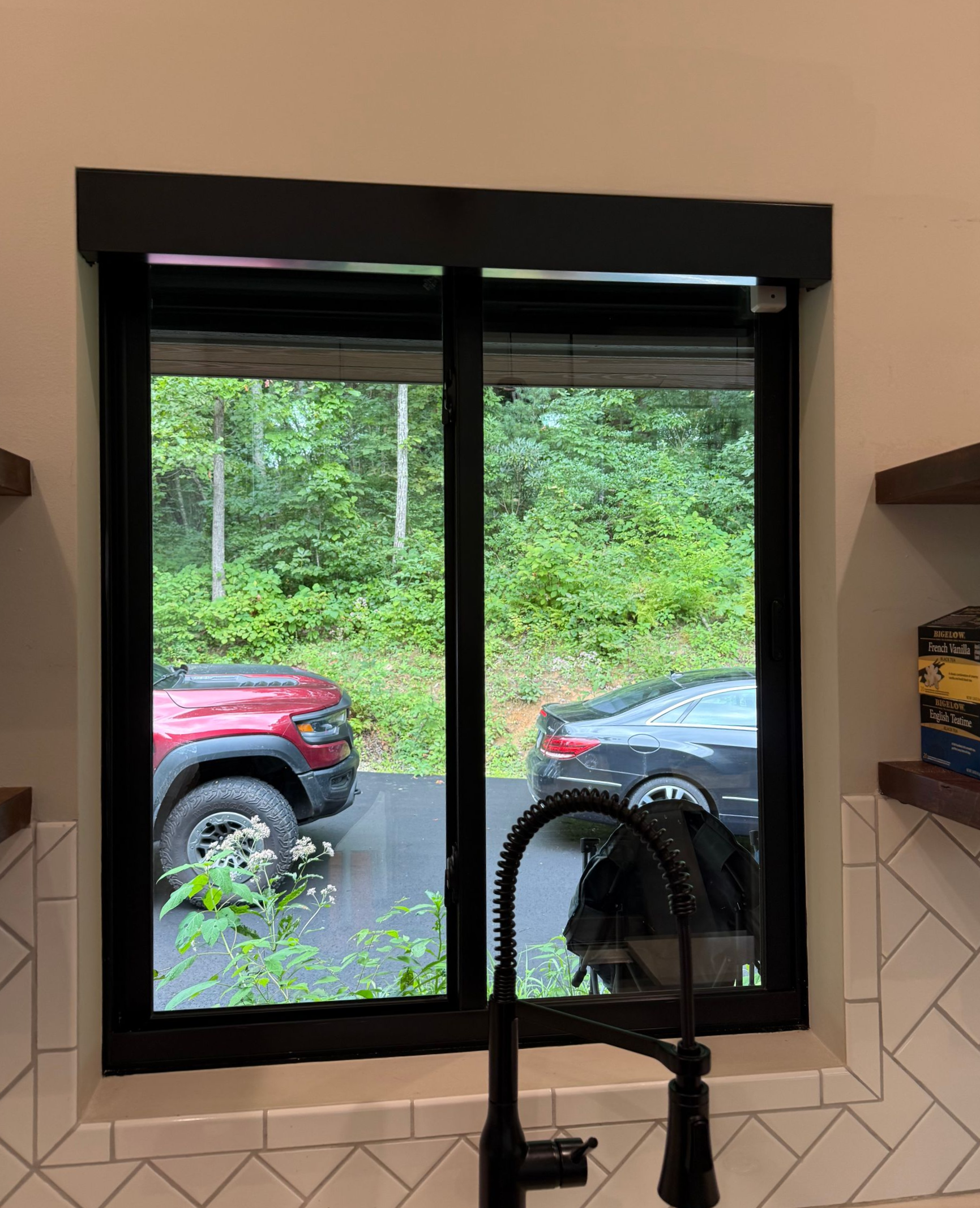 Kitchen window with black trim, showing parked cars and greenery outside behind a faucet