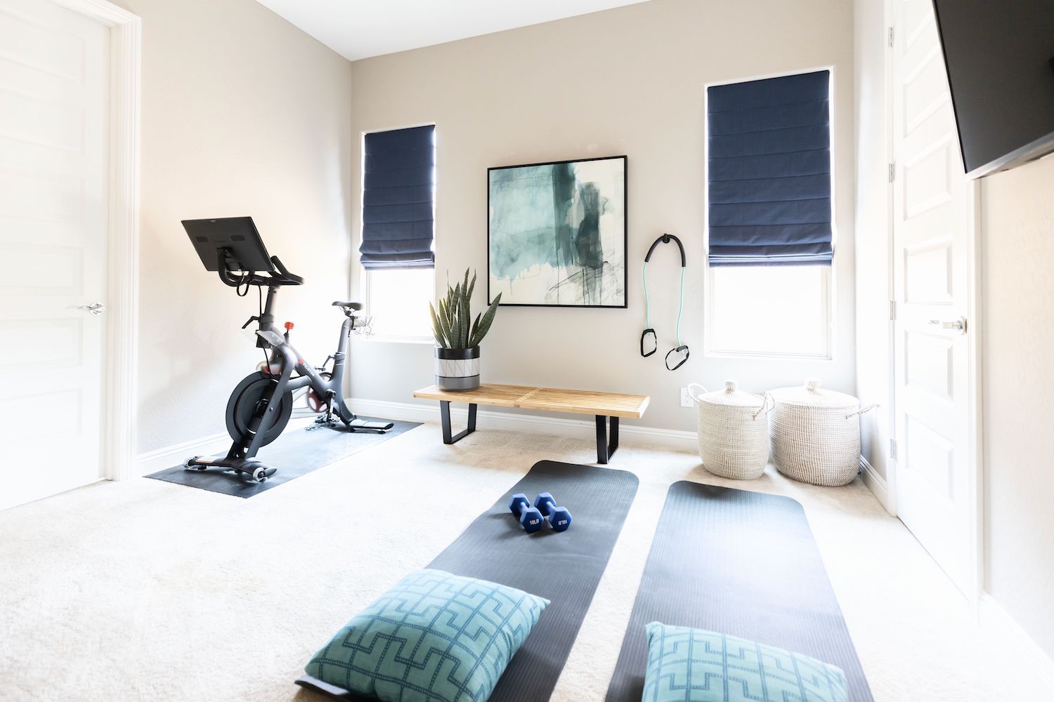 Bright home gym with exercise bike, yoga mats, dumbbells, bench, and wall art in a light room